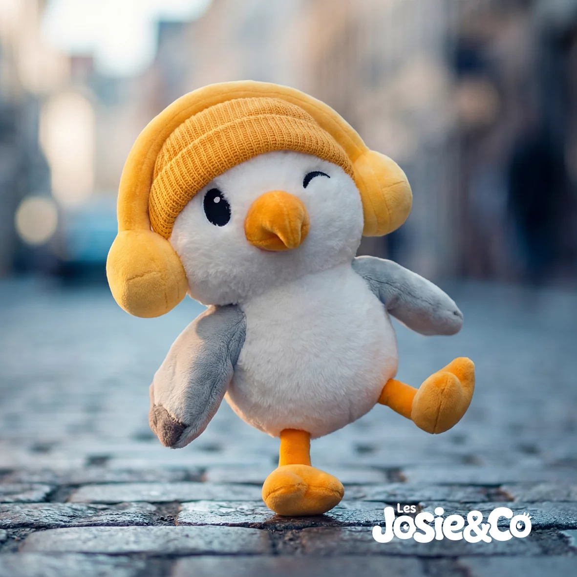 Peluche Josie la Mouette en marche sur des pavés, une patte orange levée